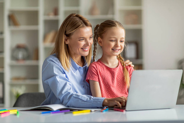 Educación en línea. Madre sonriente y su hija pequeña usando el ordenador portátil juntos en casa, cuidando a mamá ayudando al niño femenino con la tarea en la computadora, disfrutando de la enseñanza a distancia, primer plano - Foto, imagen