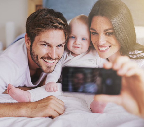 Happy family, parents and selfie of baby on bed in home for love, care and quality time together. Mother, father and newborn child smile for photograph, fun memory and happiness of bonding in bedroom. - Foto, afbeelding