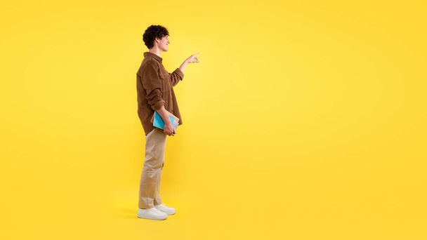 Vue latérale d'un étudiant avec des manuels pointant du doigt à l'écart de l'espace libre, faisant semblant d'appuyer sur un bouton invisible ou sonnant à la porte debout sur fond jaune studio. Panorama - Photo, image