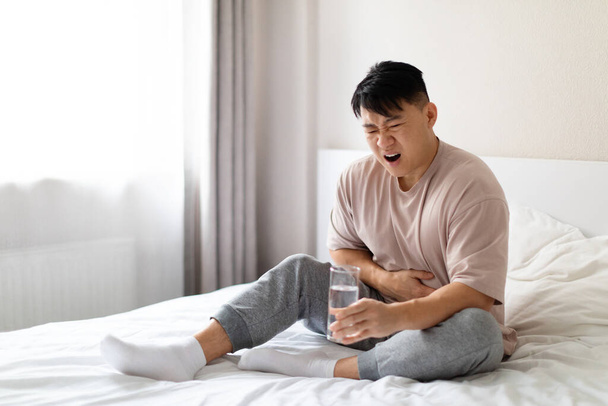 Unhappy sick middle aged chinese man sitting on bed and touching his belly, suffering from stomach pain in the morning, wearing pajamas, holding glass of water. Food poisoning, diarrhea concept - Photo, Image