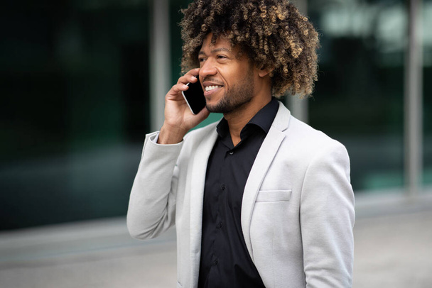 Veselý africký podnikatel středního věku mluví na smartphonu, chodí ven poblíž moderního kancelářského centra. Černošky muž podnikatel s telefonem mluvit - Fotografie, Obrázek