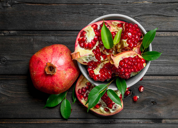 Pieces of ripe pomegranate in bowl and one ripe pomegranate with leaves. On wooden background - Foto, imagen
