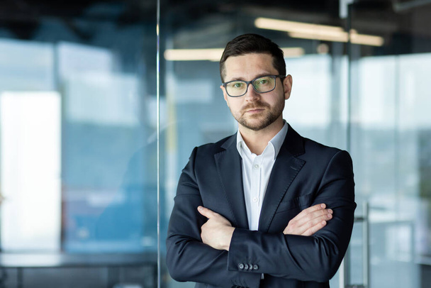 Portrait of a young businessman, a lawyer in a suit and glasses standing in the office, crossing his arms on his chest, confidently and seriously looking at the camera. - Фото, изображение