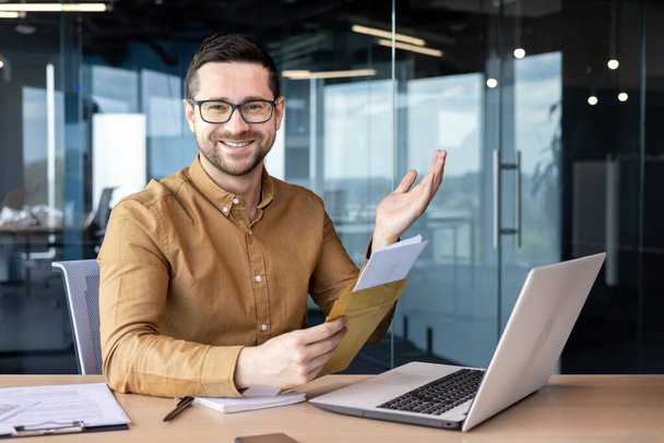 Portrait of a man sitting in the office and working on a laptop. Received and holds in his hands an envelope with a letter and documents. Smiling looking at the camera. - Foto, Imagem