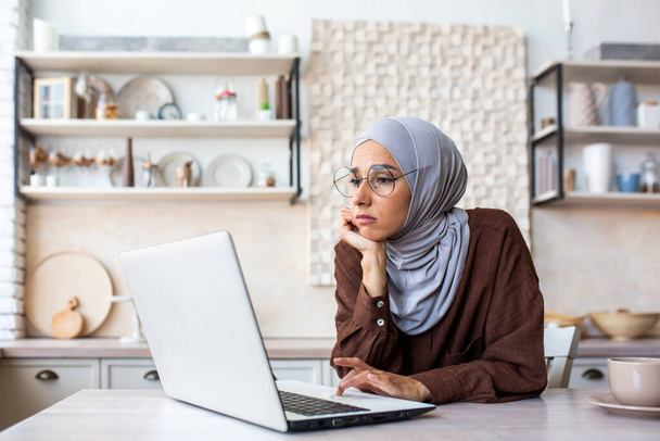 Giovane donna musulmana annoiata che indossa hijab seduto in cucina a casa, appoggiando la testa sulle mani, guardando triste e lunatico al computer portatile. - Foto, immagini