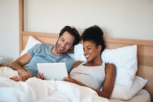 Happy couple, tablet and relax on bed for entertainment, movie or online streaming together at home. Interracial man and woman relaxing, morning or watching on technology or social media in bedroom. - Foto, Imagen