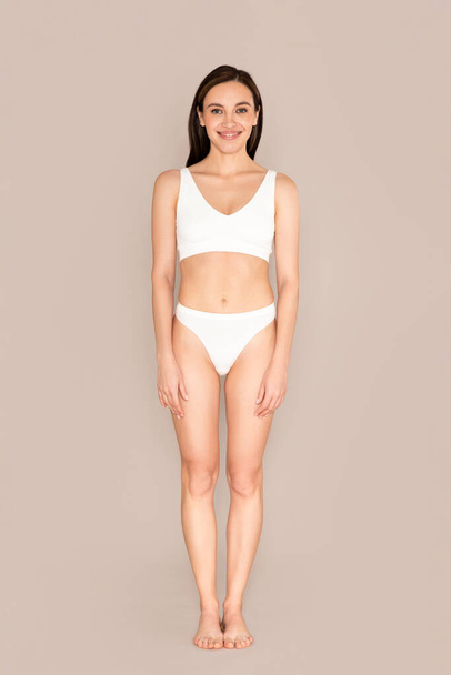 Happy pretty young slim woman with beautiful perfect body posing on beige studio background, smiling at camera, wearing white comfy underwear. Full length vertical portrait - Foto, imagen