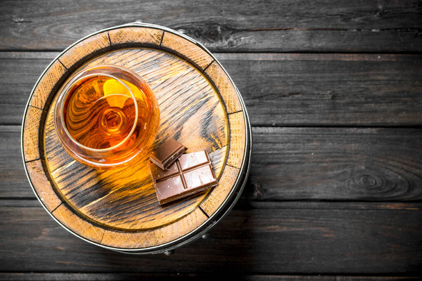 Cognac in a glass with chocolate on a barrel. On black wooden background - Zdjęcie, obraz