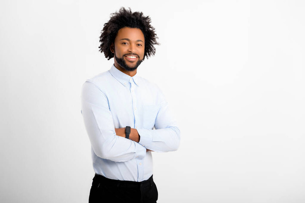 Handsome curly man looking at camera standing with arms crossed isolated on white background. African-American male employyee with curly hair in formal white shirt with folded hands - Фото, изображение