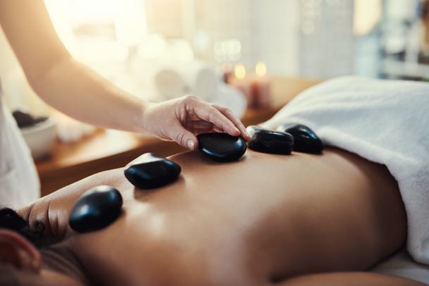 Woman, hands and rocks for back massage at spa in beauty relaxation or skincare on bed. Hand of masseuse applying hot rock or stones on female for physical therapy, zen or skin treatment at resort. - Photo, Image