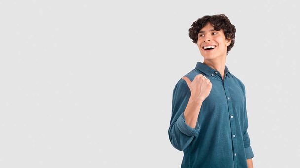 Look At This Offer. Joyful Guy Pointing Thumb Finger At Copy Space Advertising Your Text Standing On Light Gray Studio Background, Wearing Blue Shirt. I Choose This Concept. Panorama - Fotó, kép