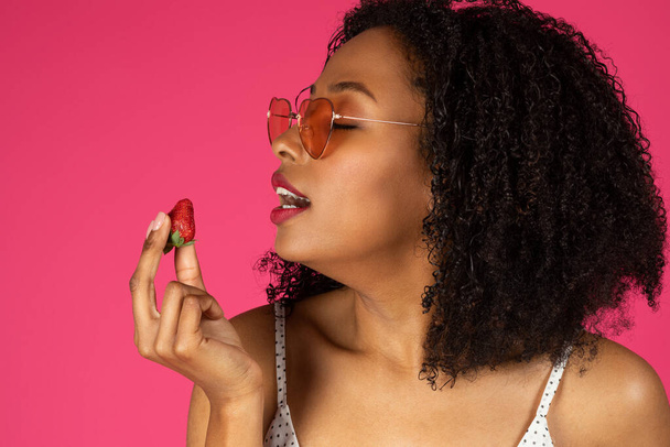 Happy young black curly lady in sunglasses eats strawberries, enjoys fruits, isolated on pink background, studio, close up. Food tasty fresh, vacation, ad and offer, beauty care - Photo, Image