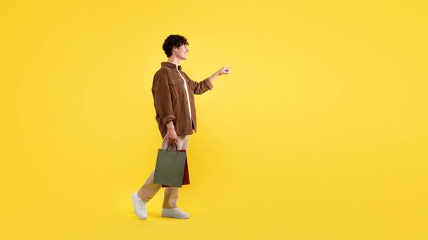 Alışveriş Teklifi Reklamı. Kağıt torbaları taşıyan ve mesaj reklamı için boş yeri işaret eden, sarı stüdyonun arkasında duran gülümseyen genç adam. Panorama - Fotoğraf, Görsel