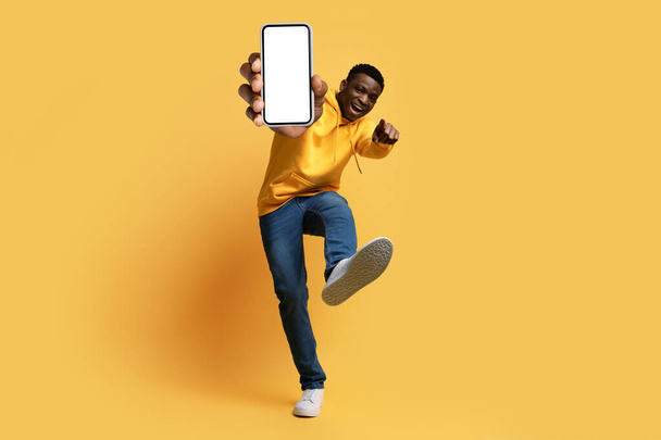 Cool χαρούμενος όμορφος νεαρός μαύρος hipster δείχνει smartphone με λευκή οθόνη, τον αντίχειρα επάνω και σόλα παπούτσι, απομονώνονται σε κίτρινο φόντο στούντιο, mockup. Ψηφιακή ζωή, σύγχρονες τεχνολογίες - Φωτογραφία, εικόνα