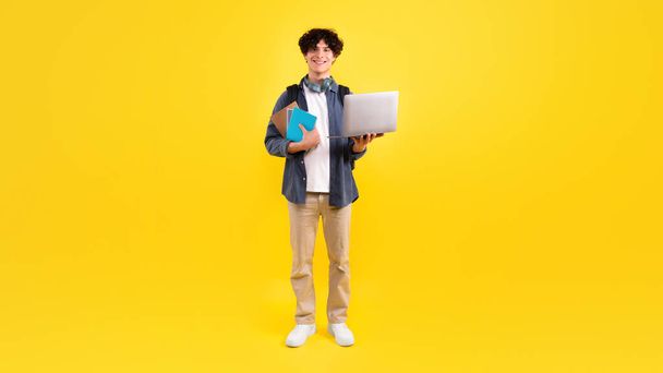 Elektronikus tanulási eszközök. Pozitív Student Guy Holding Laptop Számítógép és tankönyvek Mosolyogva a kamera, Reklám Online Oktatási Ajánlat Álló Stúdió felett Sárga Háttér. Panoráma - Fotó, kép