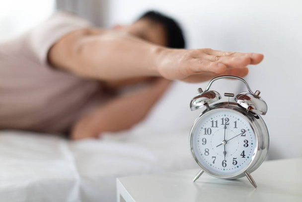 Ärgerliche Glocke, aufwachen. Unerkennbarer asiatischer Mann im Pyjama, der klingelnde Wecker abschaltet, steht auf dem Nachttisch, wacht morgens auf, verschwommener Hintergrund - Foto, Bild