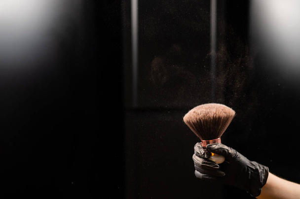 A woman shakes a large powder brush with clouds of dust on a black background. Treatment after instant tanning. - Foto, imagen