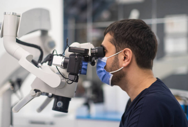 Skoncentrowany dentysta z maską medyczną na twarzy patrzący w innowacyjny mikroskop stomatologiczny sprawdzający jama pacjenta przy wizycie wysokiej jakości sprzęt w klinice stomatologicznej - Zdjęcie, obraz