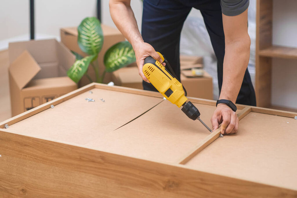 Furniture installation specialist in uniform assembling a wooden rack, attaching legs with an electric screwdriver in a residential apartment.  - Photo, Image