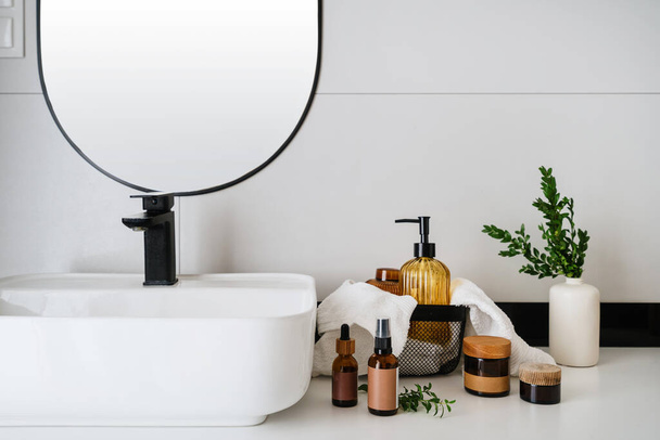 krem nawilżający w szklanym słoiku, butelka dozownika, czysty ręcznik i wazon z gałęzią roślin w pobliżu umywalki na tle ściany w nowoczesnym apartamencie, koncepcja reklamowa naturalnych kosmetyków organicznych - Zdjęcie, obraz