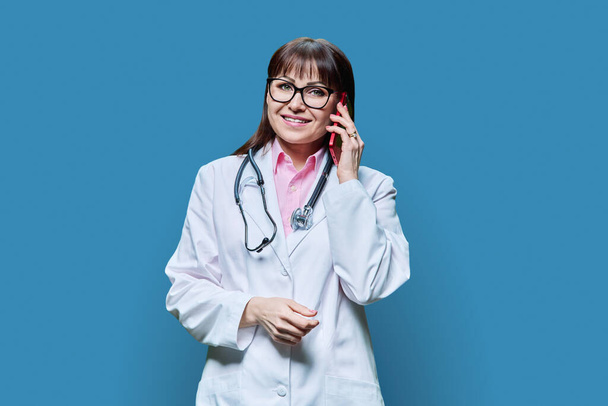 Mature female doctor in white lab coat talking on phone, on blue studio background. Medicine, service, occupation, healthcare, health care, treatment concept - Fotografie, Obrázek