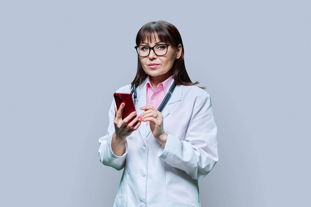 Mature female doctor in white lab coat using phone, on grey studio background. Medicine, online internet service, occupation, healthcare, health care, professional mobile apps, treatment concept - Foto, imagen