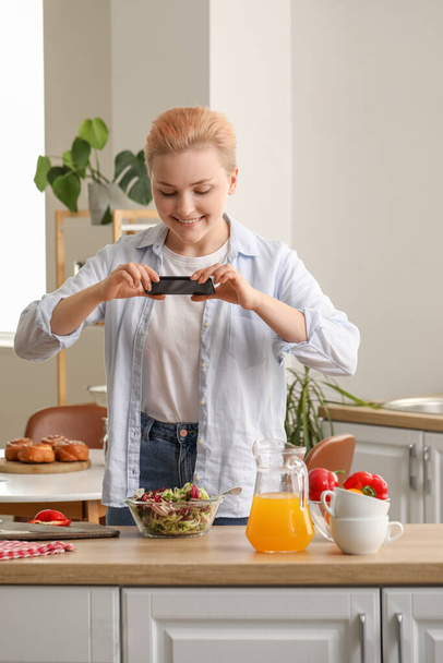 Молодая женщина с мобильным телефоном фотографирует овощной салат на кухне - Фото, изображение