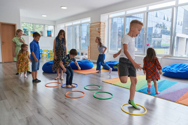 Small nursery school children with female teacher on floor indoors in classroom, doing exercise. Jumping over hula hoop circles track on the floor - Foto, Imagem