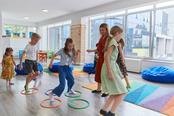 Kleine Kindergartenkinder mit einer Lehrerin auf dem Fußboden im Klassenzimmer, die Sport treiben. Springen über Hula-Hoop-Reifen kreist Bahn auf dem Boden - Foto, Bild