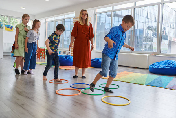 Small nursery school children with female teacher on floor indoors in classroom, doing exercise. Jumping over hula hoop circles track on the floor - Foto, immagini
