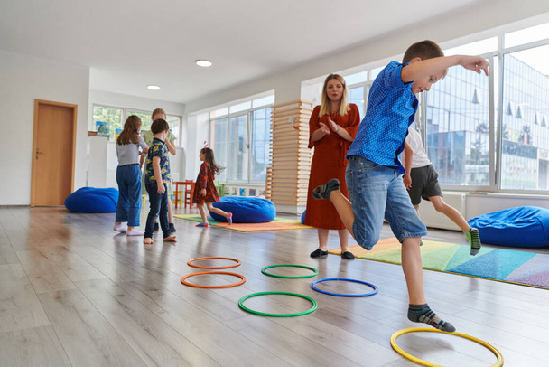 Small nursery school children with female teacher on floor indoors in classroom, doing exercise. Jumping over hula hoop circles track on the floor - Photo, Image