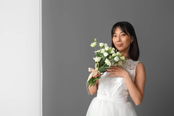 Beautiful young Asian bride with bouquet of eustoma flowers on grey background - Fotó, kép