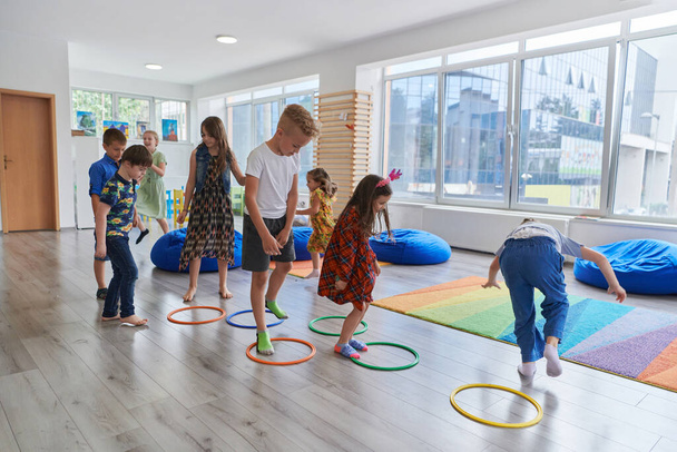 Small nursery school children with female teacher on floor indoors in classroom, doing exercise. Jumping over hula hoop circles track on the floor - Foto, afbeelding