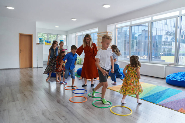 Small nursery school children with female teacher on floor indoors in classroom, doing exercise. Jumping over hula hoop circles track on the floor - Foto, afbeelding