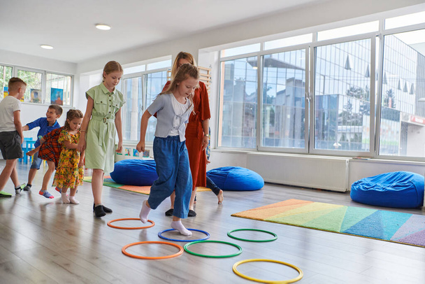Small nursery school children with female teacher on floor indoors in classroom, doing exercise. Jumping over hula hoop circles track on the floor - Fotoğraf, Görsel