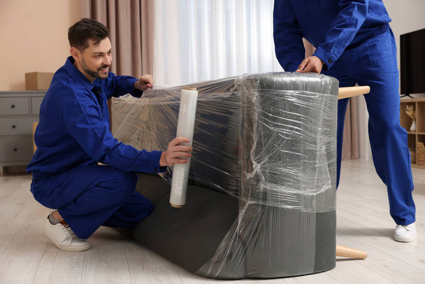 Male movers with stretch film wrapping sofa in new house - Zdjęcie, obraz