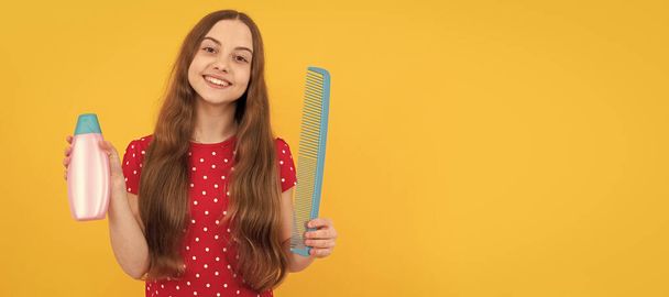happy kid long hair showing conditioner and comb on yellow background, haircare. Kid girl hair care, horizontal poster. Banner header with copy space - Zdjęcie, obraz