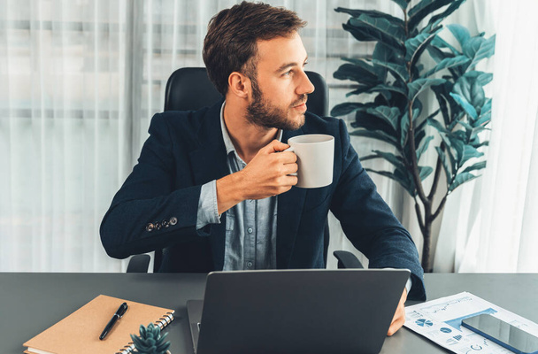 Empresário trabalhando em seu laptop e bebendo café em sua mesa no espaço de trabalho. Executivo inteligente fazendo coffee break depois de pesquisar dados financeiros e plano de marketing em seu laptop corporativo. Entidade - Foto, Imagem