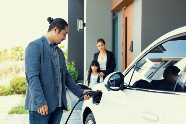 Progressive young parents and daughter with electric vehicle and home charging station. Green and clean energy from electric vehicles for healthy environment. Eco power from renewable source at home. - Foto, Imagen