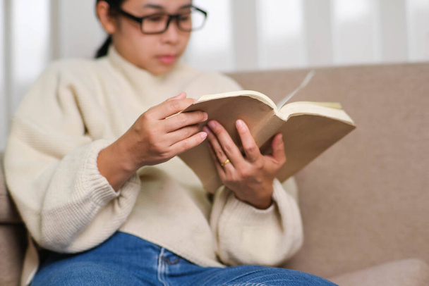 Счастливая азиатка сидит на диване и читает книгу в помещении. Портрет женщины в белом свитере, читающей книгу на диване дома. Стиль жизни, свободное время и отдых. - Фото, изображение