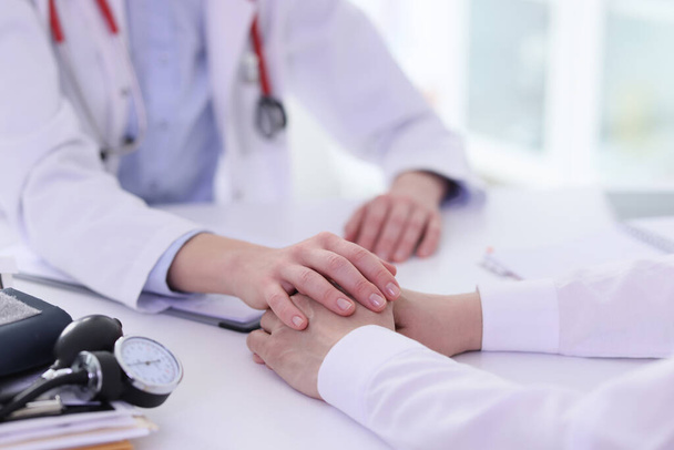 Γιατρός κρατά τα χέρια του αρσενικού πελάτη κατά τη διάρκεια της διαβούλευσης κάθεται στο ελαφρύ τραπέζι στο γραφείο κλινική. Ειδικός υποστηρίζει τον ασθενή με περίπλοκη ασθένεια - Φωτογραφία, εικόνα