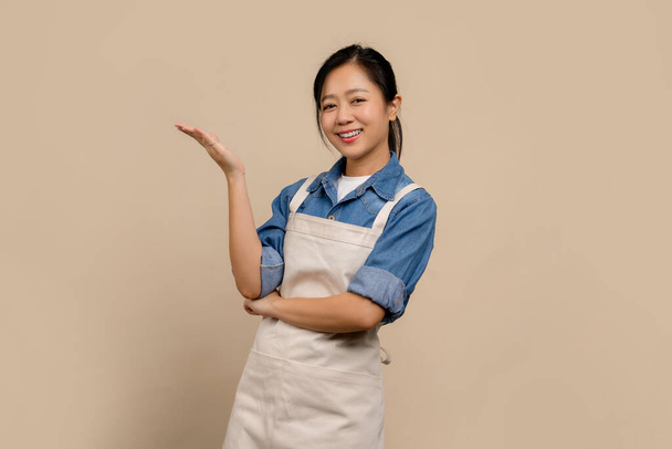 Portrait of cheerful Asian woman wearing apron on cream background - Photo, image