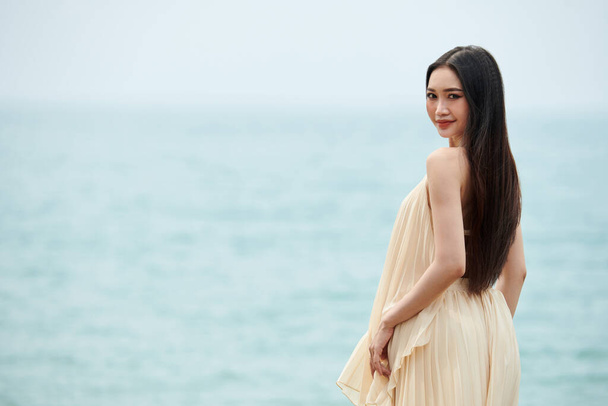 Молодая женщина с длинными волосами, стоящая на пляже и поворачивающаяся к камере - Фото, изображение