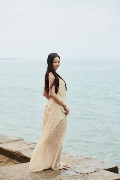 Young Asian woman in long flowing dress standing on beach and smiling at camera - Φωτογραφία, εικόνα