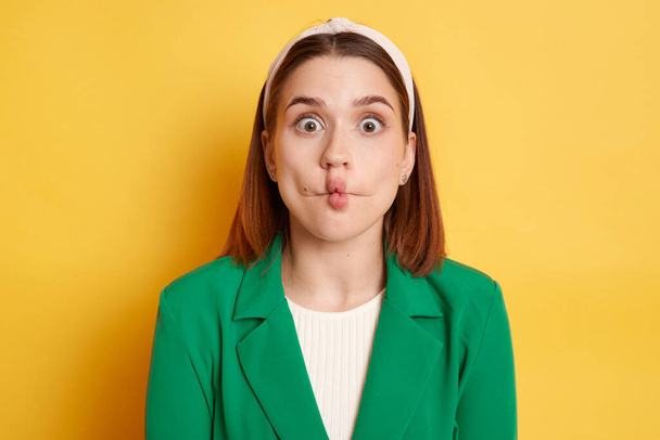Funny optimistic woman wearing green jacket making fish lips grimacing having fun looking at camera with big eyes posing isolated over yellow background. - Foto, Imagen