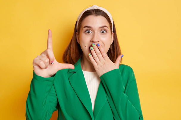 Shocked amazed woman wearing green jacket posing isolated over yellow background showing looser gesture looking at camera with surprised eyes covering mouth with palm. - Фото, зображення