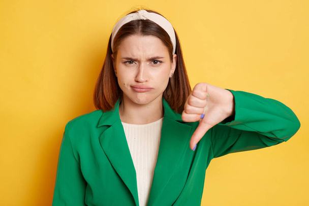 Displeased sad woman wearing green jacket grimacing showing thumb down demonstrates dislike gesture posing isolated over yellow background. - Fotó, kép