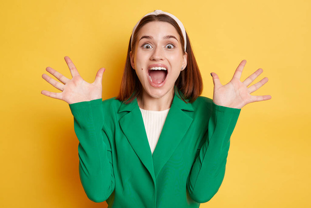 Extremely happy surprised woman wearing green jacket posing isolated over yellow background raised her arms screaming with happiness excited emotions. - Foto, Imagen