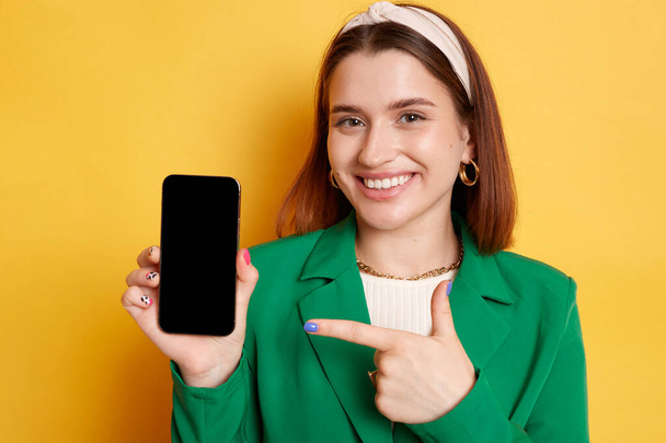 Pretty smiling woman wearing green jacket posing isolated over yellow background pointing at smartphone with empty display empty space for advertisement. - Foto, Imagen