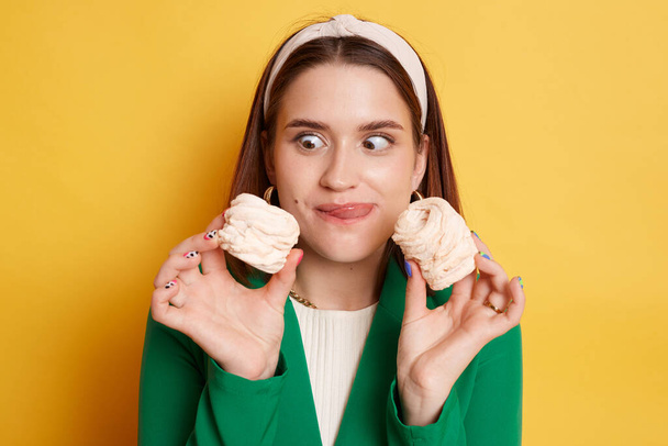 Crazy funny woman wearing green jacket posing isolated over yellow background looking at marshmallow with big eyes tongue out wants to eat sugary confectionery. - Photo, Image